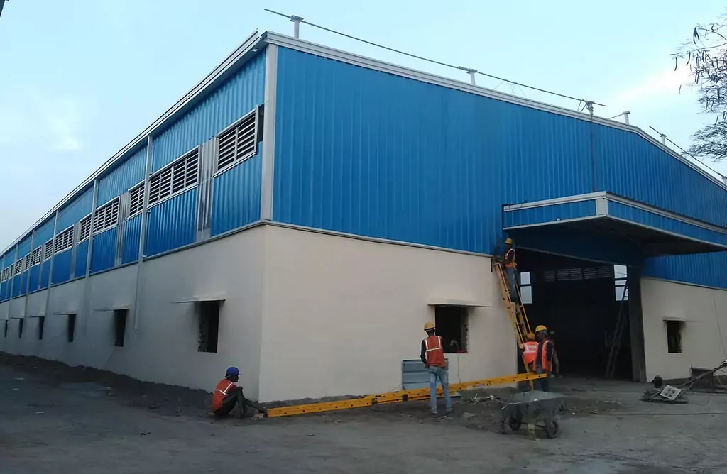 A blue and white corrugated metal warehouse under construction, featuring workers finishing the lower masonry walls, depicting the setup phase of Factory Sheds built by Steel City as experienced Industrial Shed Manufacturers in Pakistan.