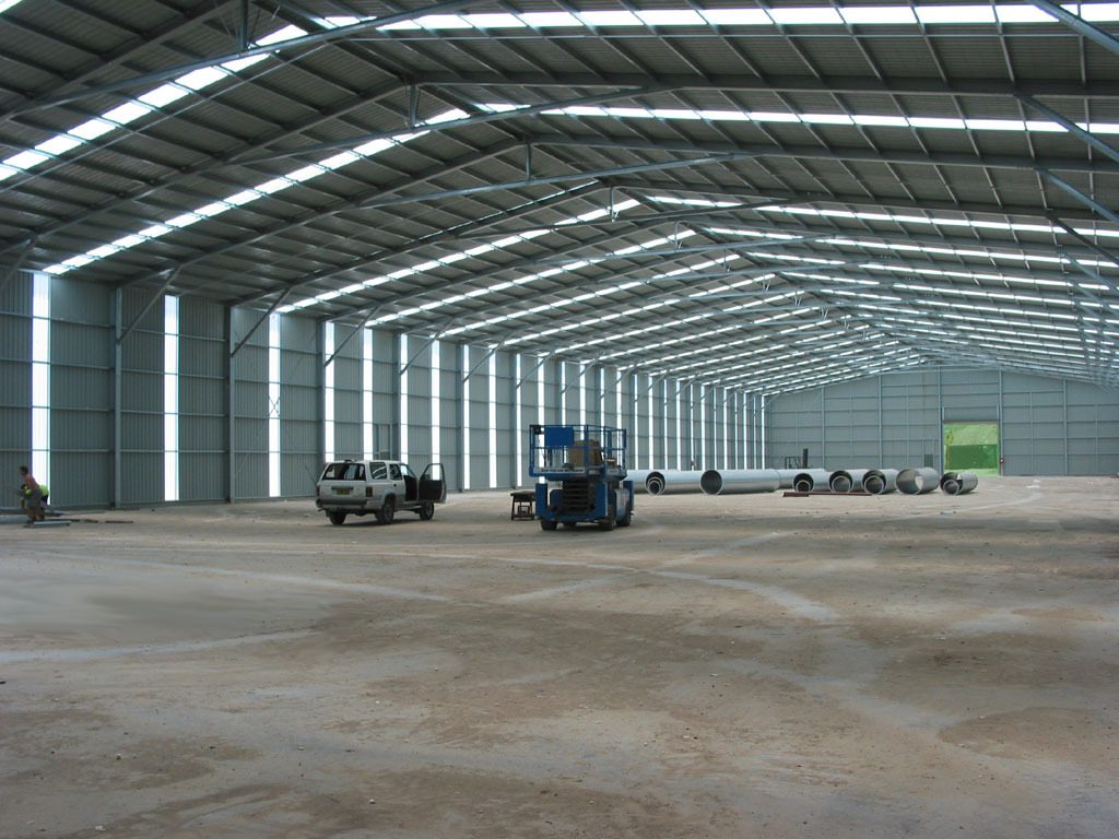 A wide interior view of a large steel truss warehouse under construction, featuring a scissor lift, utility vehicle, and structural steel pipes. This large-span design is typical of heavy-duty Factory Sheds designed by specialized Industrial Shed Manufacturers.
