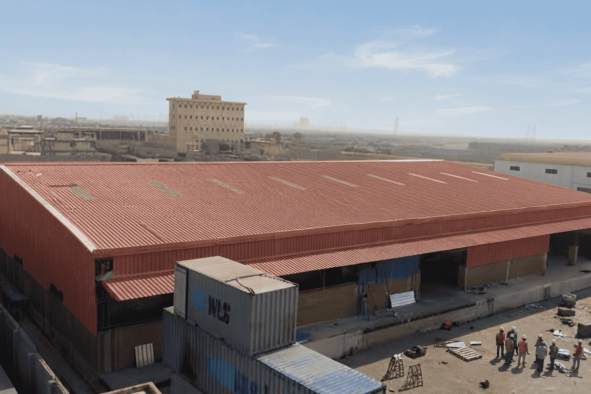 High-angle view of an industrial site featuring a long steel shed warehouse with a red corrugated roof and open bays. Stacked blue shipping containers sit in the foreground, while a small group of workers in hard hats stands in a dusty yard under a clear sky.